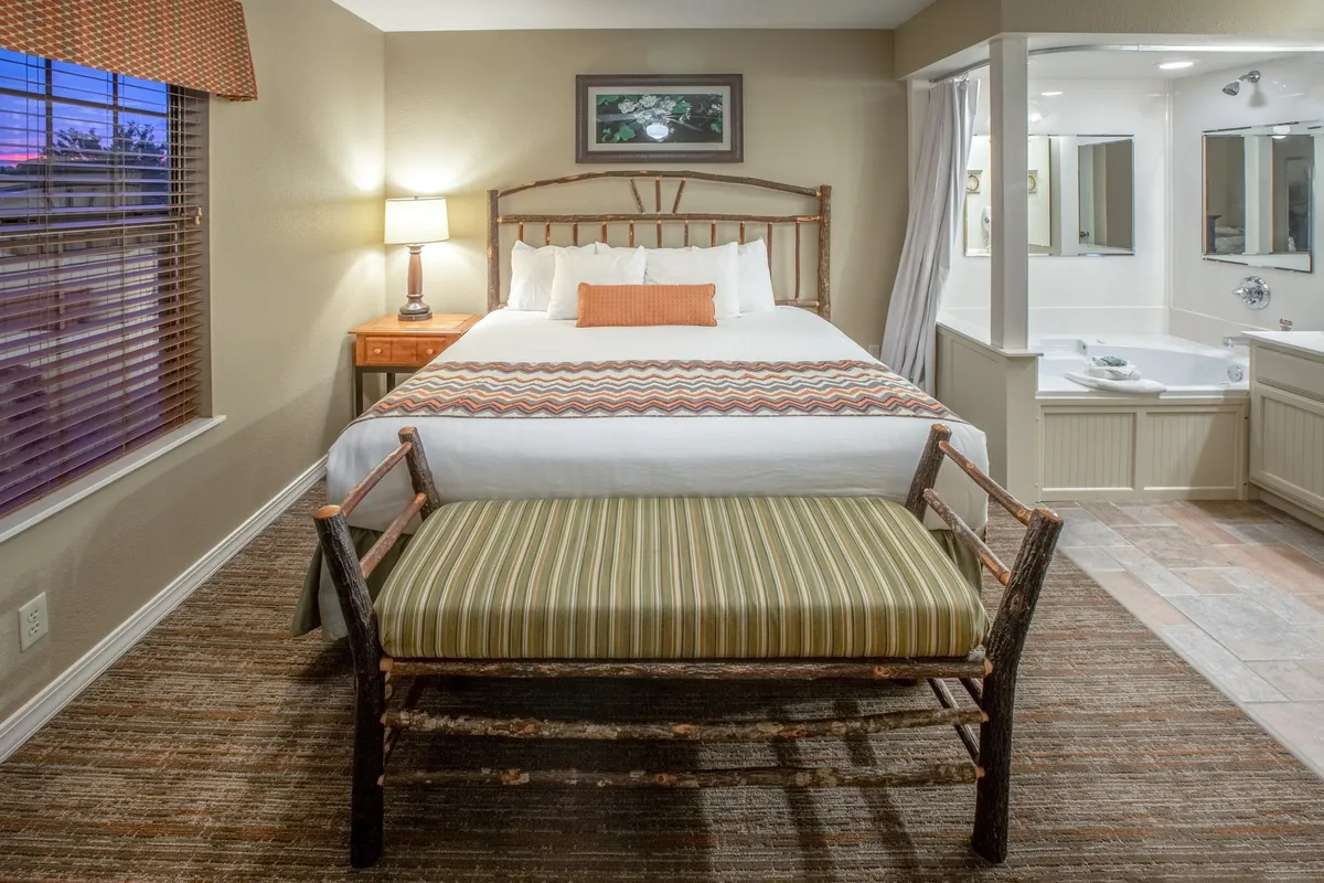 The image showcases a serene and inviting master bedroom, featuring a plush bed with decorative pillows, a cozy bench at the foot, a softly glowing bedside lamp, and a glimpse of a modern, spa-like bathroom—creating a tranquil sanctuary for relaxation.