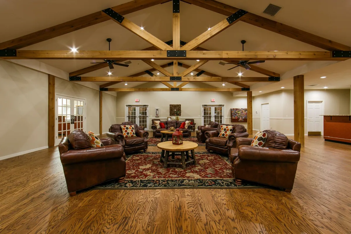 A cozy living room scene featuring plush leather sofas arranged around a round coffee table atop a decorative rug. Wooden beams add architectural warmth overhead, and natural light streams in from large windows, illuminating the inviting space. The setting is clearly designed for social gatherings, relaxation, or intimate conversations, and the overall mood is warm and welcoming.