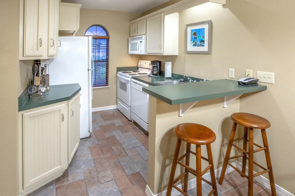 This image showcases a bright, welcoming kitchen interior with a spacious countertop, bar stools, modern appliances, and a sunlit window view. The scene is set for both cooking and socializing, capturing moments like friends enjoying a casual brunch and family preparing meals together. The overall atmosphere is lively yet comfortable, emphasizing the convenience and communal feel of the kitchen and adjacent dining area.